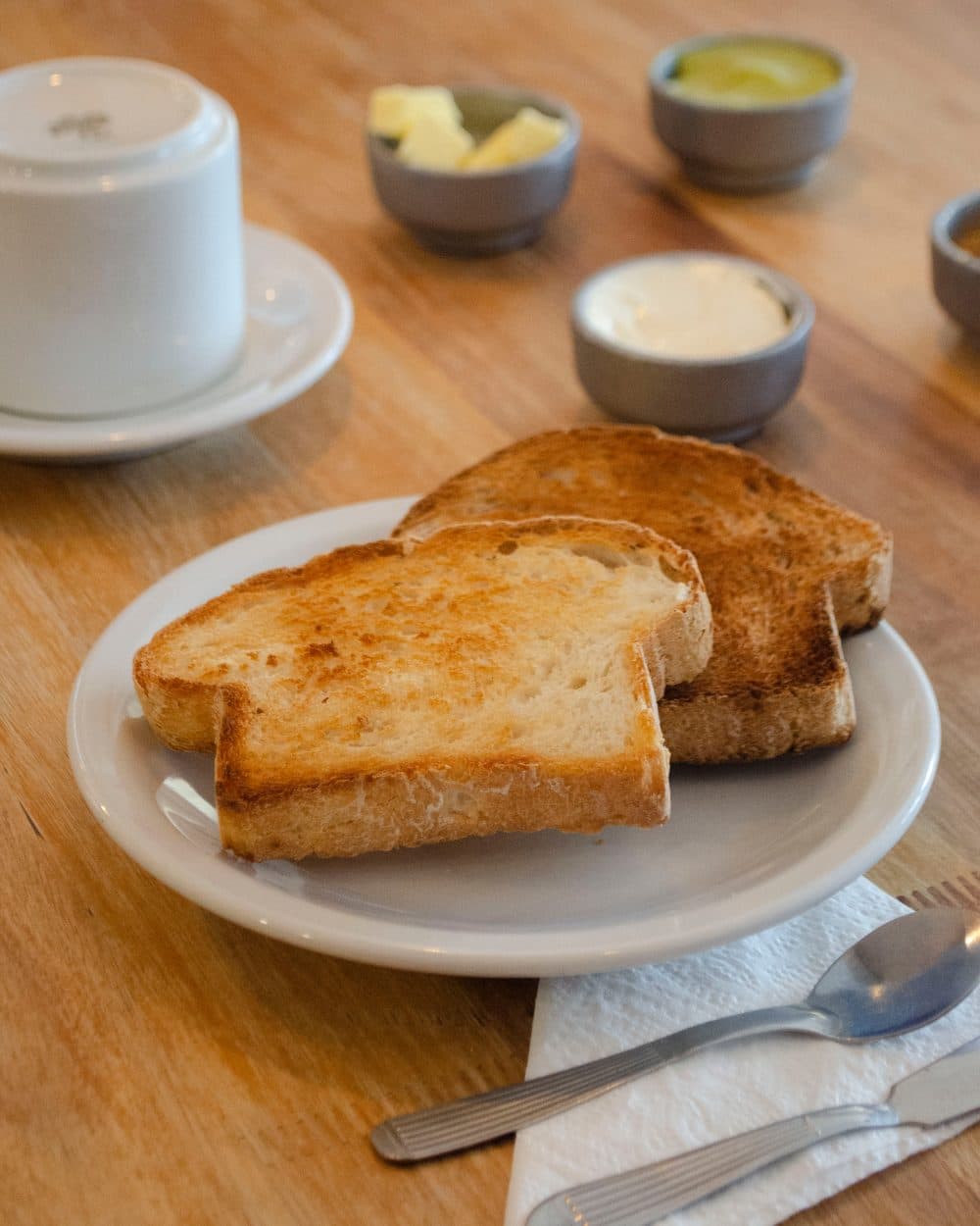 Mesa de desayuno servida: dos tostadas en un plato blanco, una taza de café, manteca y cuencos con mermelada sobre una mesa de madera